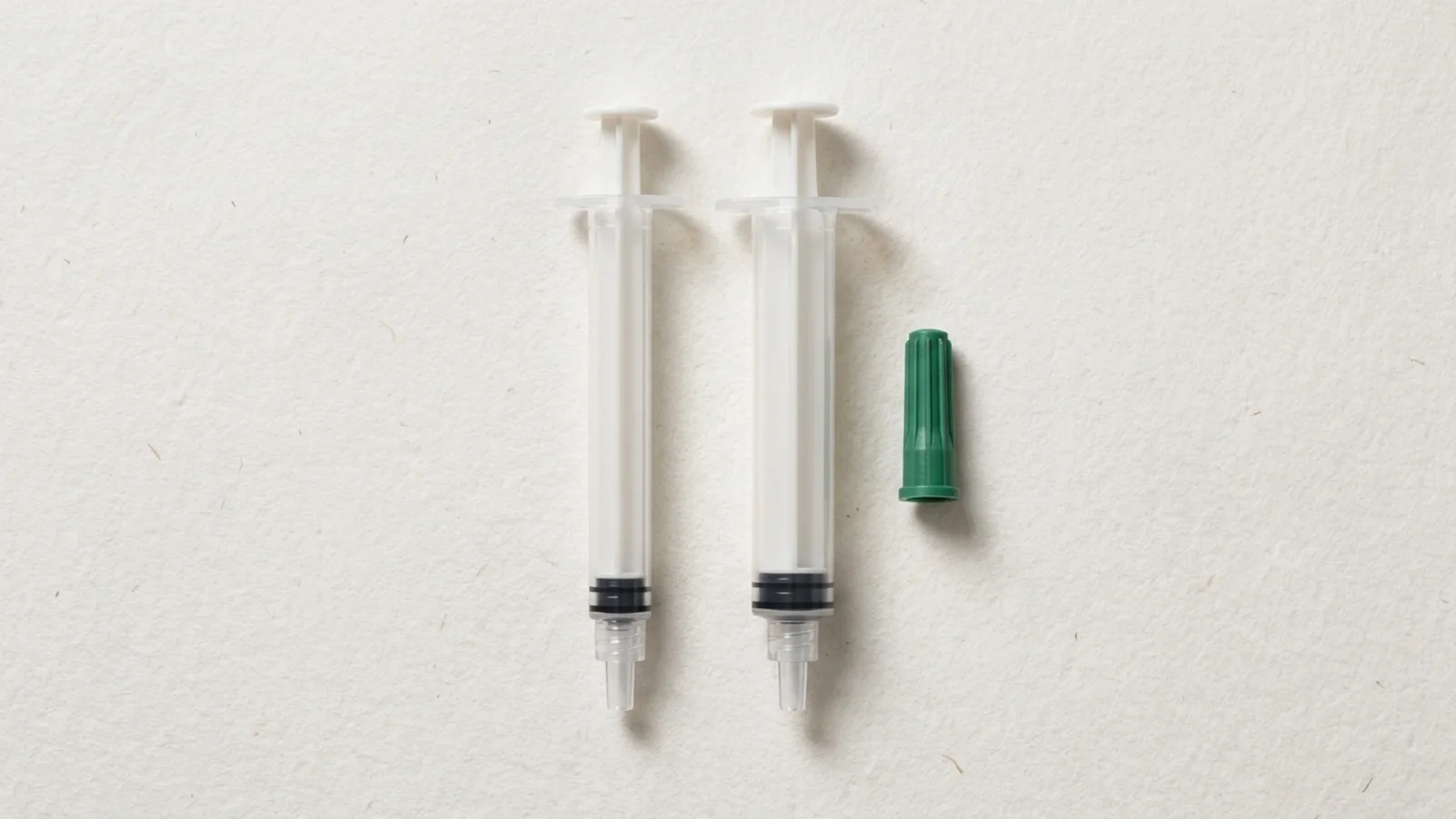 Top-down catalog still life of two empty arterial blood-gas syringes with translucent barrels and a detached green tip cap on an off-white background