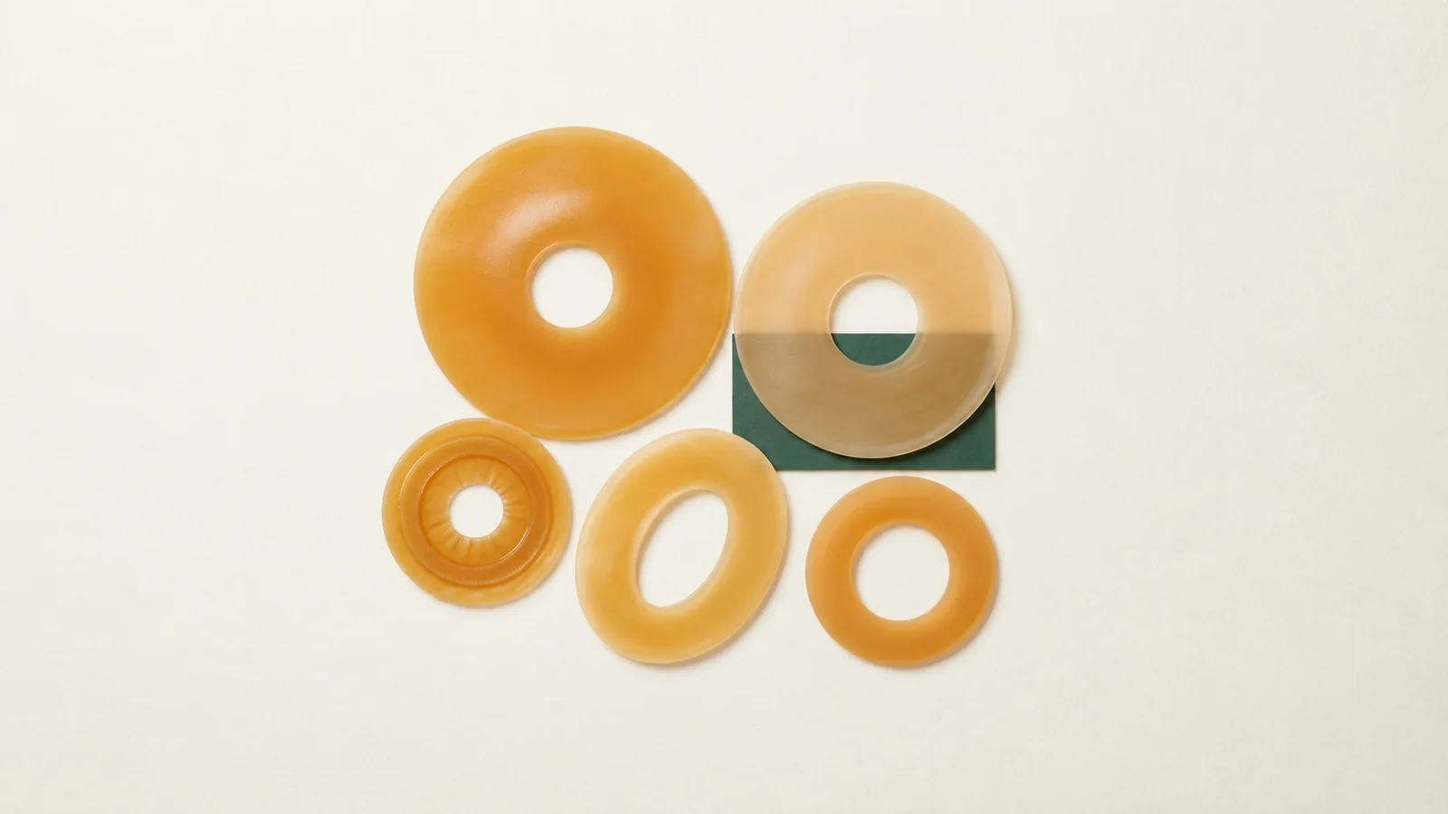 Illustrative top-down still life of five unbranded amber ostomy barrier rings, including one oval, grouped on an off-white surface with a green backing accent.
