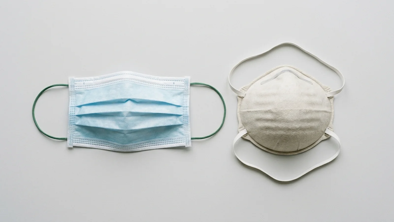 Text-free editorial still life comparing a pleated surgical mask and an unmarked filtering-facepiece respirator on pale gray