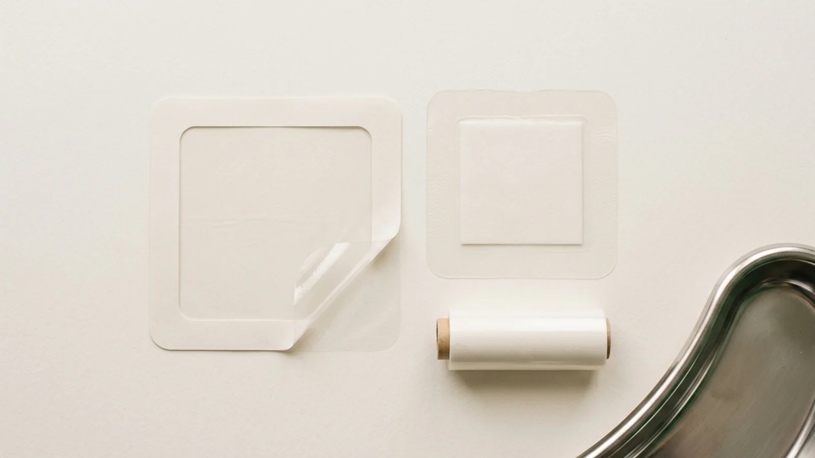 Transparent film dressings in frame-style, bordered island, and roll formats arranged on an off-white surface with a stainless steel tray edge for specification comparison.